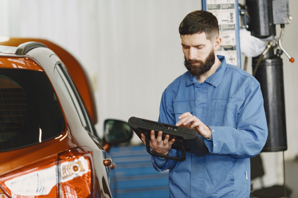 Technician in coveralls running an automotive diagnostic check beside a car